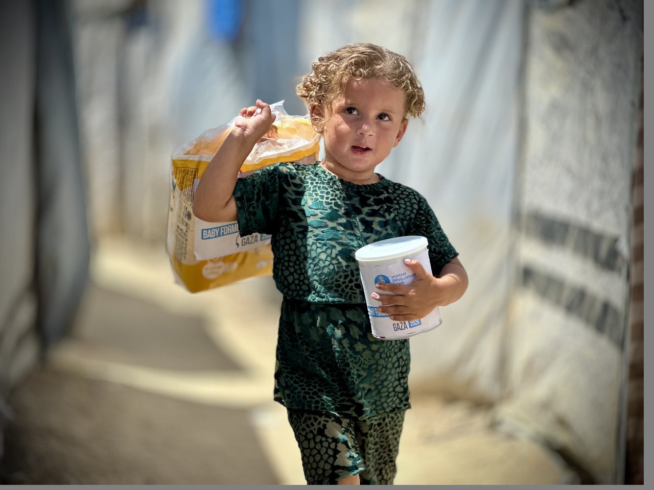 Child in a Gaza displacement camp carrying a yellow HDF baby formula bag and a can of infant formula
