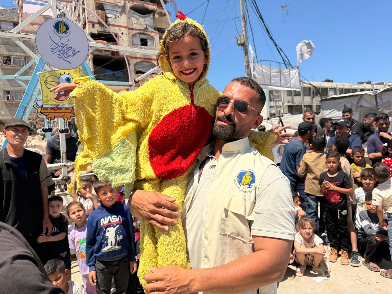 HDF field worker in a branded vest lifting a smiling child at a Gaza community gathering