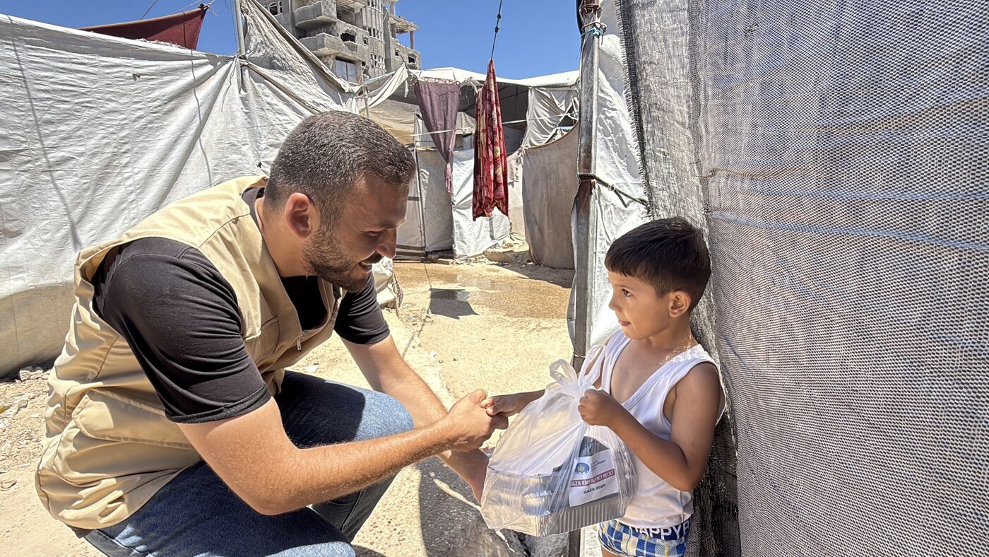 An HDF staff member crouching to hand food to a boy beside a shelter tent in Gaza.